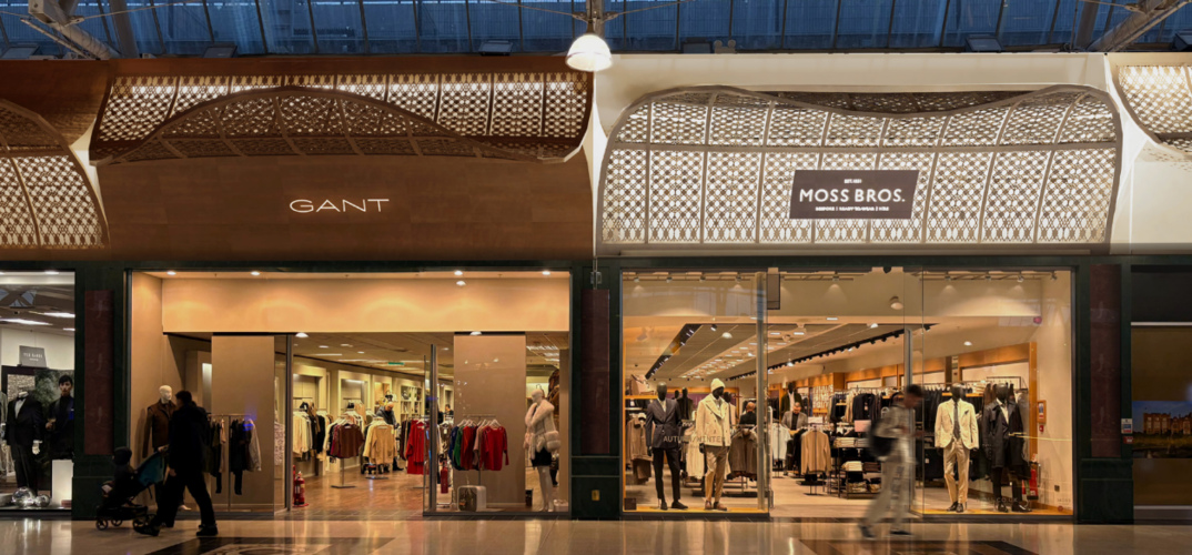 Shop fronts inside an indoor shopping complex. The store fronts show sleek metal detailing above the store names, giving a classy mesh pattern with light and metal.